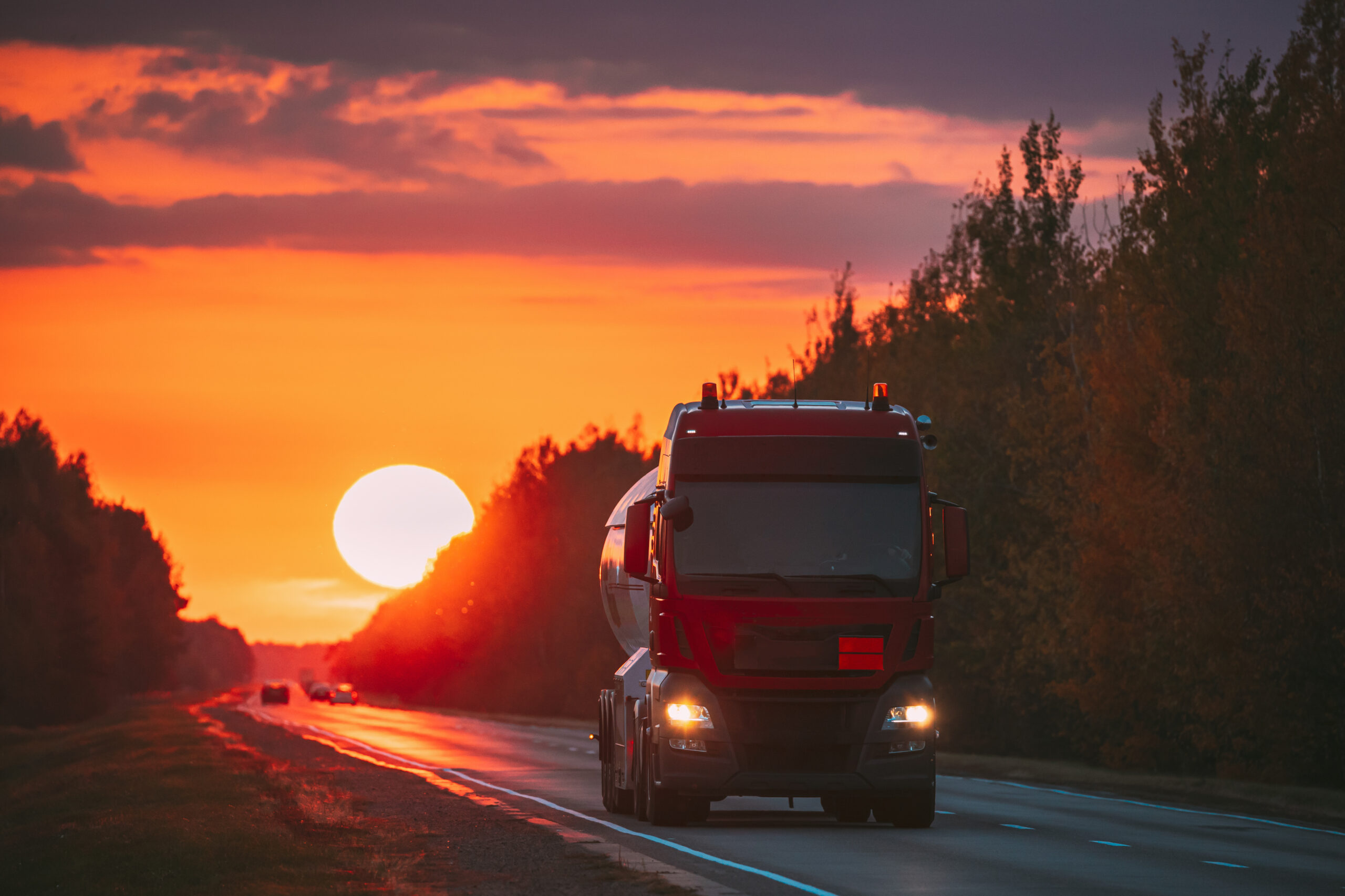 Red Truck Or Tractor Unit, Prime Mover, Traction Unit In Motion On Road, Freeway. Asphalt Motorway Highway Against Background Of Big Sunset Sun. Business Transportation And Trucking Industry
